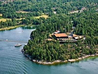 Flygfoto över en ö med täta träd, en stor byggnad och en brygga. En liten båt närmar sig kajen, omgiven av vatten och grönska.