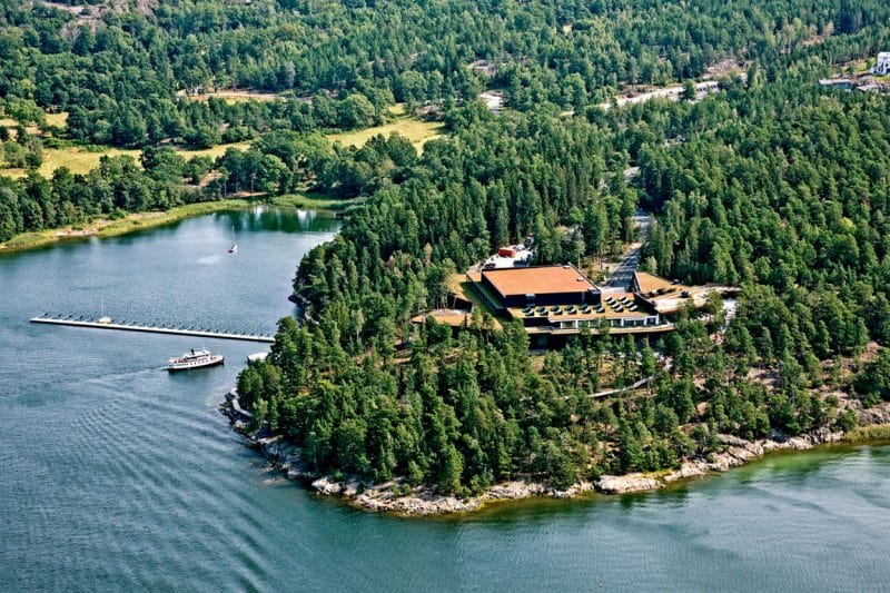 Flygfoto över en ö med täta träd, en stor byggnad och en brygga. En liten båt närmar sig kajen, omgiven av vatten och grönska.