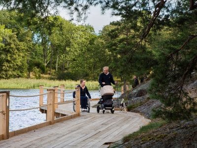 Två personer färdas längs en träspång vid sjön med barnvagn och rullstol, omgiven av grönska och träd.