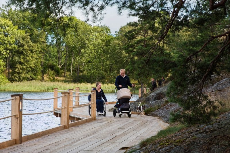 Två personer färdas längs en träspång vid sjön med barnvagn och rullstol, omgiven av grönska och träd.