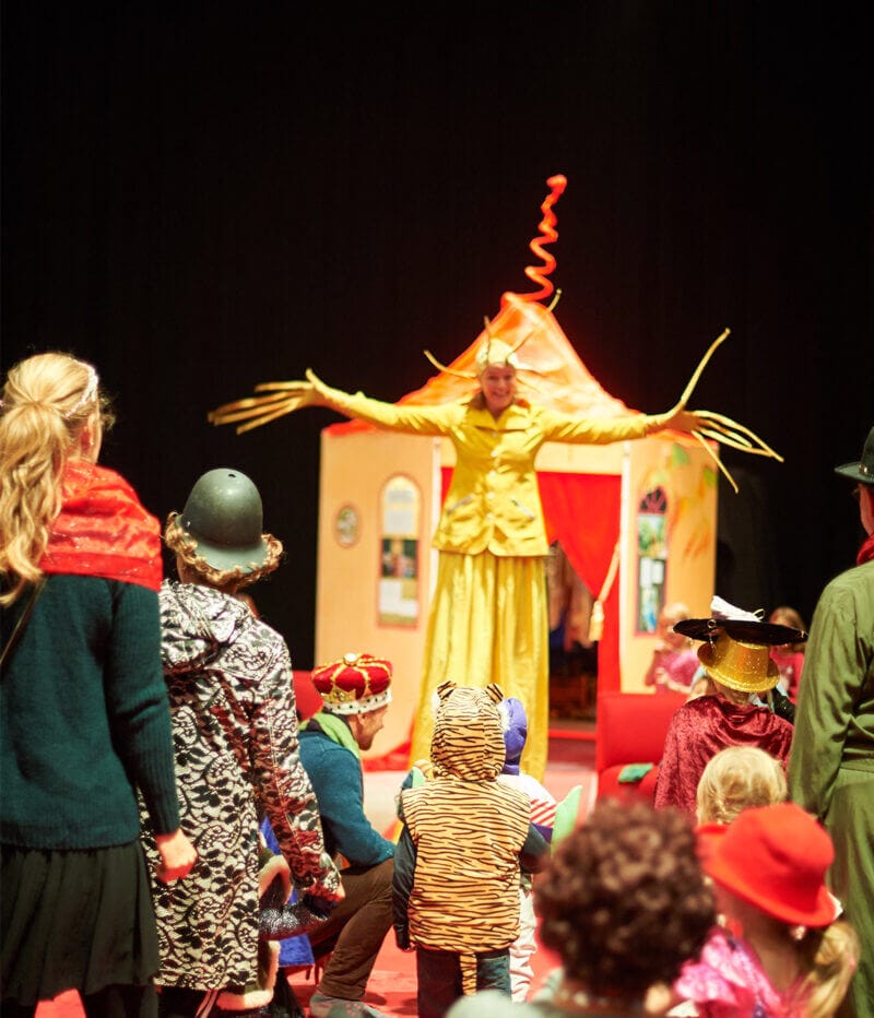 kvinna klädd i gul dräkt med jättelånga fingrar öppnar upp famnen för att välkomna barn och vuxna till ett färgglatt lekland