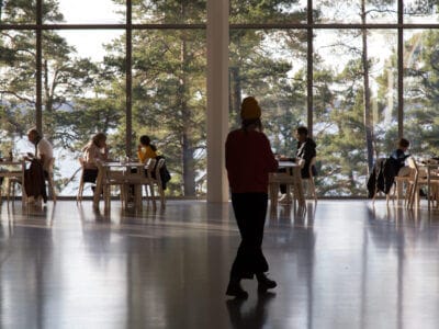 En person går över en stor hall med ett blankt golv och upplever Artipelagkvällars fridfulla atmosfär. I bakgrunden sitter folk vid bord vid stora fönster med utsikt över ett skogsområde och njuter av Artipelag kvällstids unika charm.