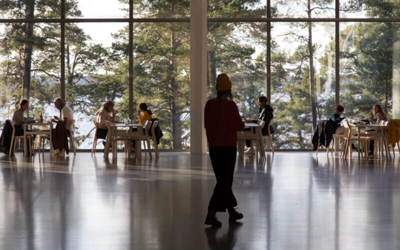 En person går över en stor hall med ett blankt golv och upplever Artipelagkvällars fridfulla atmosfär. I bakgrunden sitter folk vid bord vid stora fönster med utsikt över ett skogsområde och njuter av Artipelag kvällstids unika charm.
