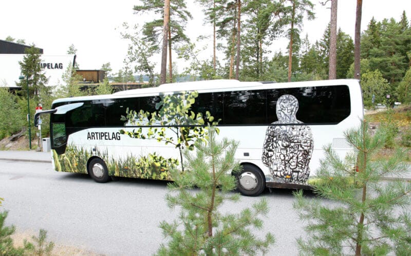 En vit buss med "ARTIPELAG"-text och en naturinspirerad abstrakt design, parkerad på en trädkantad väg, inbjuder till att uppleva Artipelagkvällar – upplev Artipelag kvällstid.