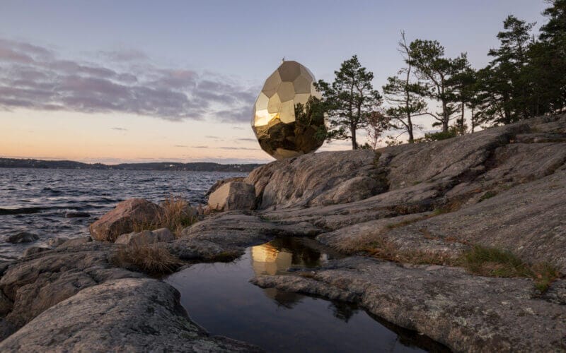 En stor reflekterande guldäggskulptur sitter i stenig terräng vid en vattenmassa, med träd i bakgrunden under en halvmolnig himmel, och erbjuder en fängslande upplevelse som påminner om Artipelagkvällar – Upplev Artipelag kvällstid.