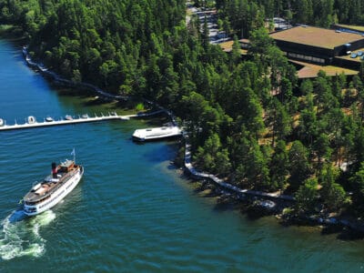 Flygbild på Artipelag. Bilden visar en passagerarbåt som strax ska lägga till vid ångbåtsbryggan. I bild syns även hela Artipelags byggnad ovan ifrån, den kringliggande tallskogen och träspången som slingrar sig längs med kustlinjen.