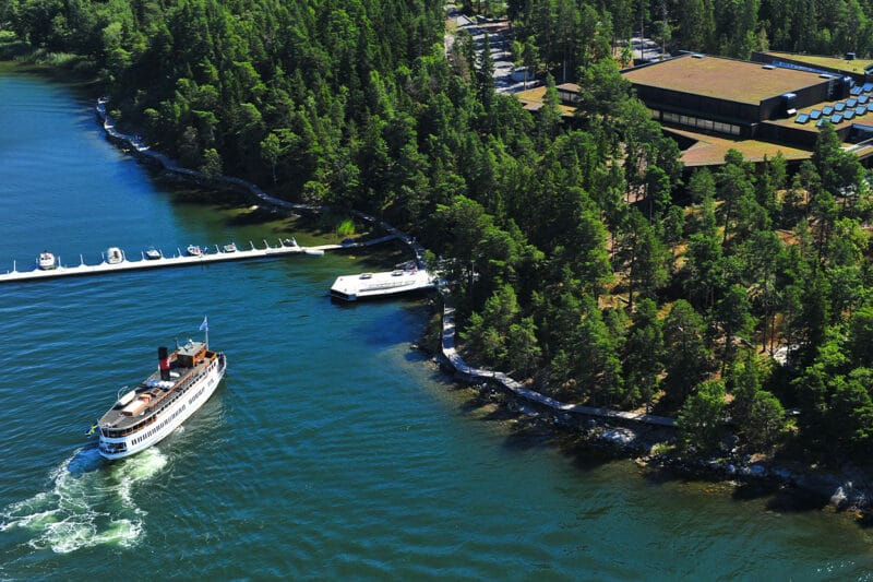 Flygbild på Artipelag. Bilden visar en passagerarbåt som strax ska lägga till vid ångbåtsbryggan. I bild syns även hela Artipelags byggnad ovan ifrån, den kringliggande tallskogen och träspången som slingrar sig längs med kustlinjen.