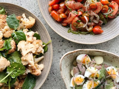 Bild på tre rätter ovanifrån. Den ena rätten föreställer en sallad innehållandes rostad blomkål. Den andra rätten är en sallad med tomater och tunt skivad lök. Den tredje rätten är en lerpottasill med matjesill, ägg, potatis och dill.