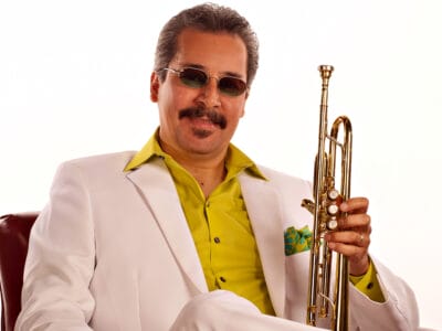 En man i vit kostym och gul skjorta sitter i en läderfåtölj och håller i en trumpet.