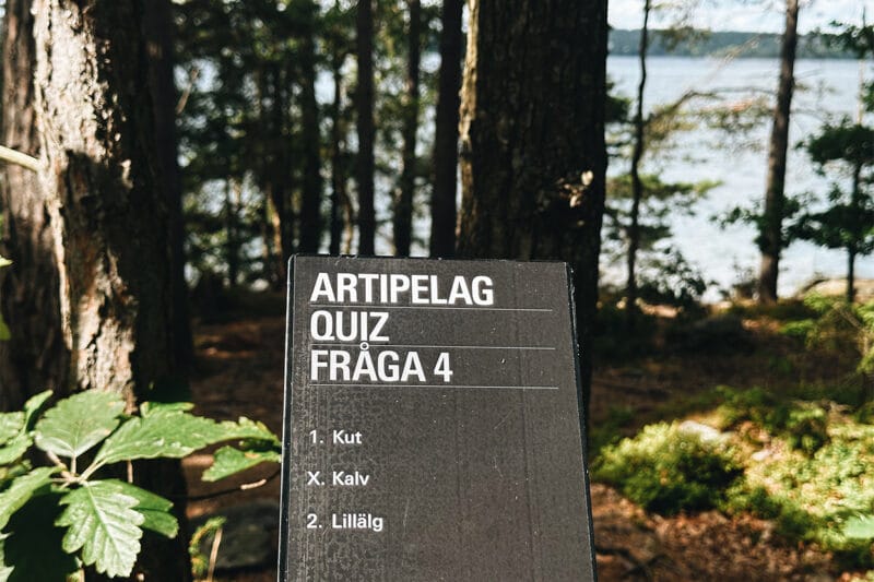 Bilden föreställer en skylt med en fråga från Artipelags tipspromenad. I bakgrunden skymtar skog och vatten.