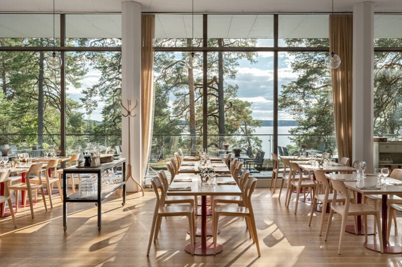Bilden förställer Artipelag Restaurang i solljus. På bilden syns dukade bord på rad framför ett panoramafönster med utsikt över vattnet och talltopparna.