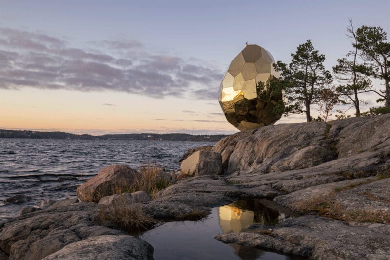 Bilden föreställer skulpturen Solar Egg i skymning. Skulpturen i form av ett gyllene ägg står på en klippa vid vattnet, solen har precis gått ner i horisonten.