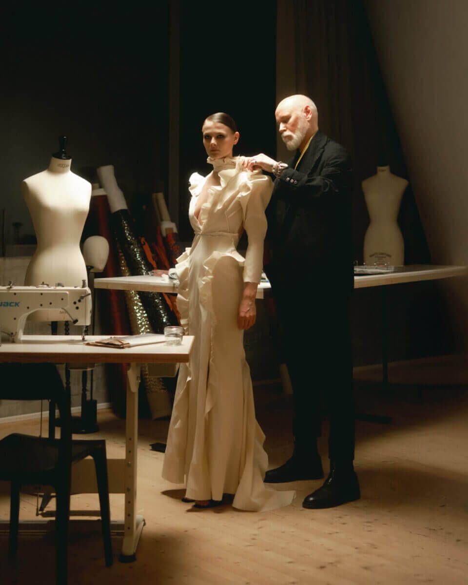 I Lars Wallin Atelier anpassar en designer en lång, vit klänning på en modell, omgiven av skyltdockor, bord och symaskiner. Studion firar 35 år av modehistorier och surrar av kreativ energi.