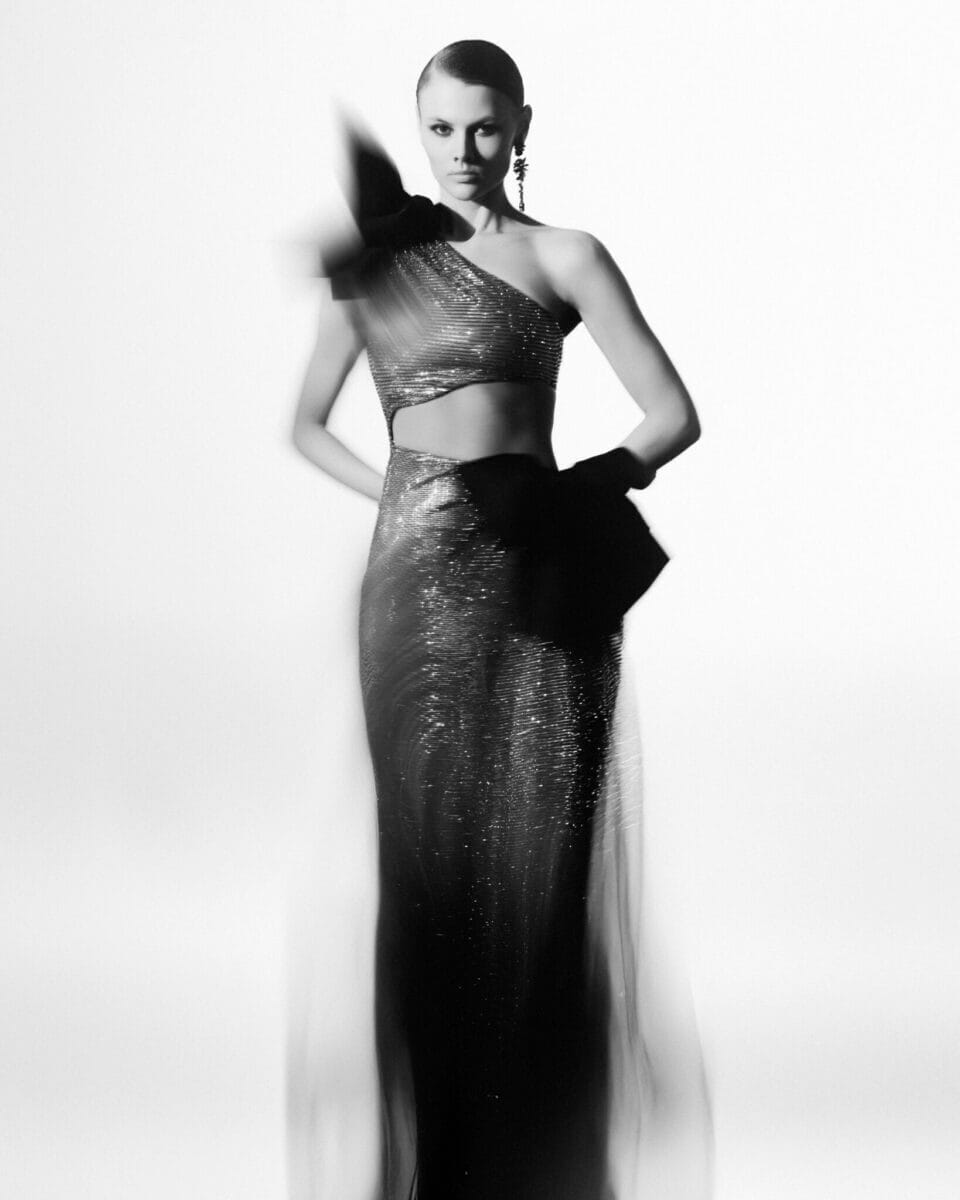 En person är elegant poserad i en skimrande klänning med en axel, prydd med en stor båge, händerna säkert placerade på höfterna. Kanterna på klänningen suddas ut mjukt i den här svartvita bilden – en hyllning av stilen från Lars Wallin Atelier – 35 Years of Fashion Stories.