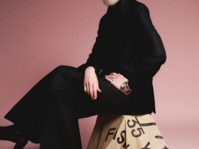 En skäggig man i svart outfit, modedesignern Lars Wallin, sitter på en tygklädd pall mot en rosa bakgrund och återspeglar essensen av 35 Years of Fashion Stories.