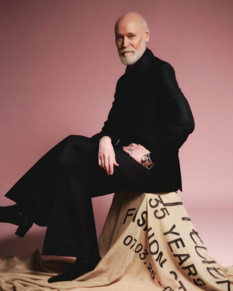 En skäggig man i svart outfit, modedesignern Lars Wallin, sitter på en tygklädd pall mot en rosa bakgrund och återspeglar essensen av 35 Years of Fashion Stories.