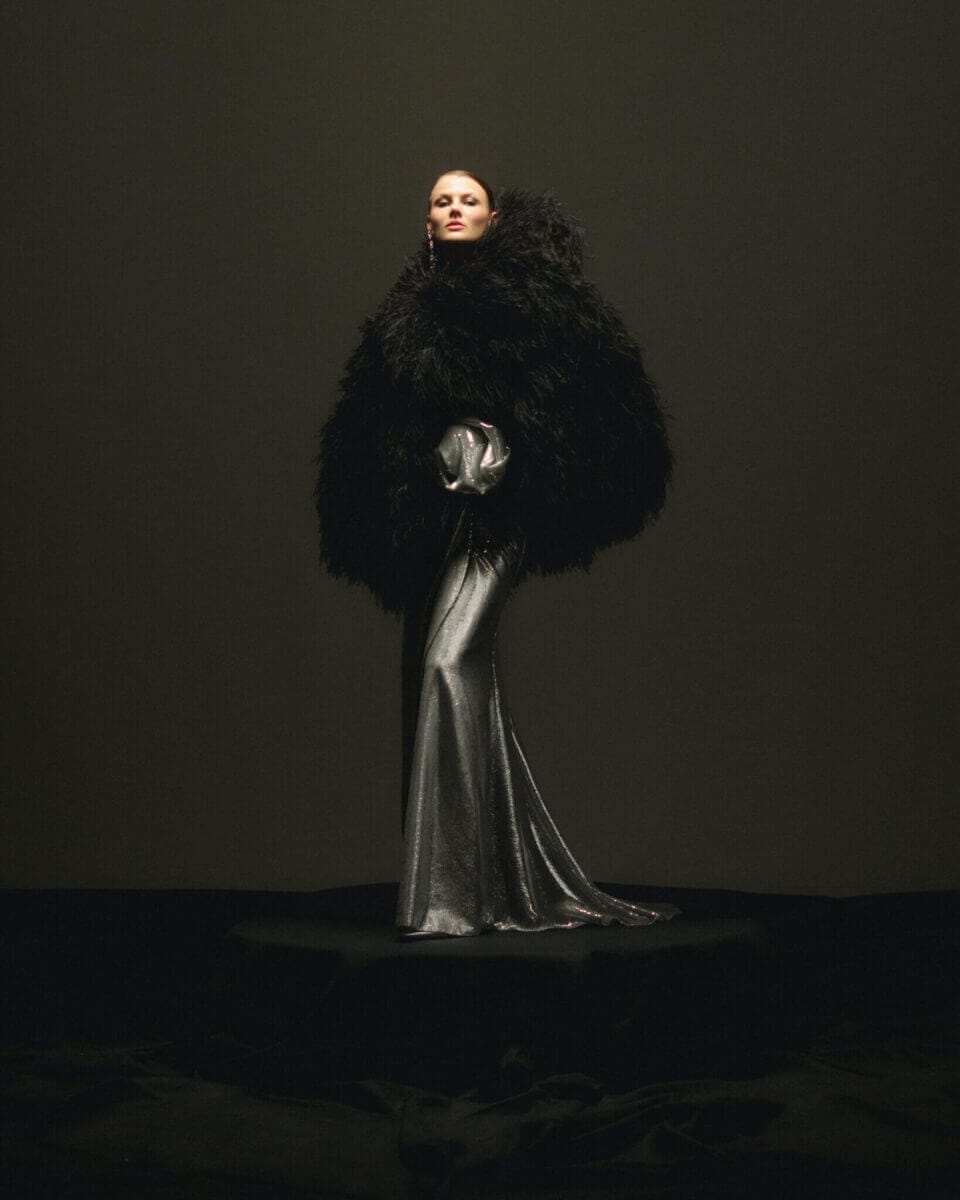 En person är elegant utsmyckad i en dramatisk, stor svart fjäderkappa över en metallisk silverklänning, mot en mörk bakgrund. Denna fantastiska ensemble fångar essensen av Lars Wallin Atelier – 35 Years of Fashion Stories