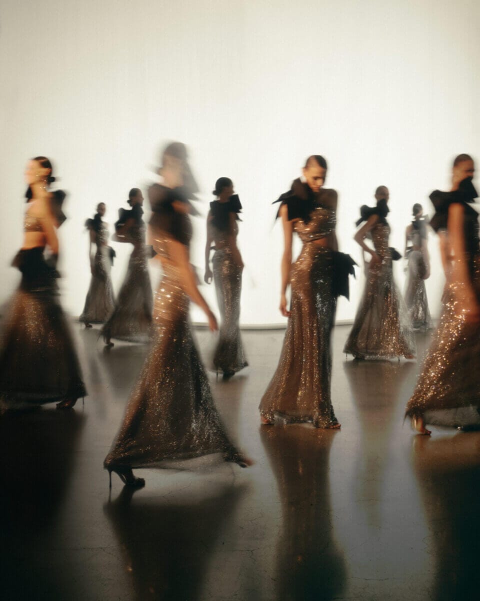 En grupp modeller i långa, skimrande klänningar går och poserar i ett mjukt upplyst utrymme, och deras rörelseoskärpa fångar den dynamiska andan hos Lars Wallin Atelier – 35 Years of Fashion Stories.