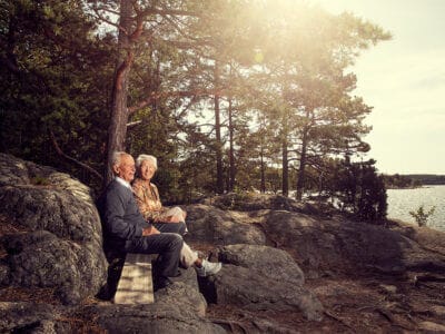 An elderly couple sits on a wooden bench by the rocky shore, surrounded by trees with sunlight streaming through, much like a scene you might find around Artipelag.