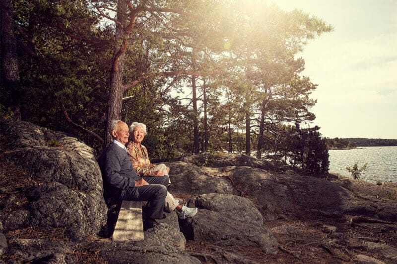 An elderly couple sits on a wooden bench by the rocky shore, surrounded by trees with sunlight streaming through, much like a scene you might find around Artipelag.