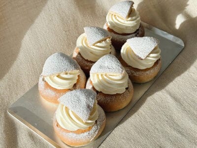 En bricka med sex nygräddade semlor placeras på en beige tygbakgrund och bjuder in till Bådan Café & Konditori på Värmdö i Stockholms skärgård för en härlig upplevelse.