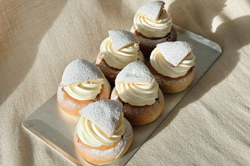 En bricka med sex nygräddade semlor placeras på en beige tygbakgrund och bjuder in till Bådan Café & Konditori på Värmdö i Stockholms skärgård för en härlig upplevelse.