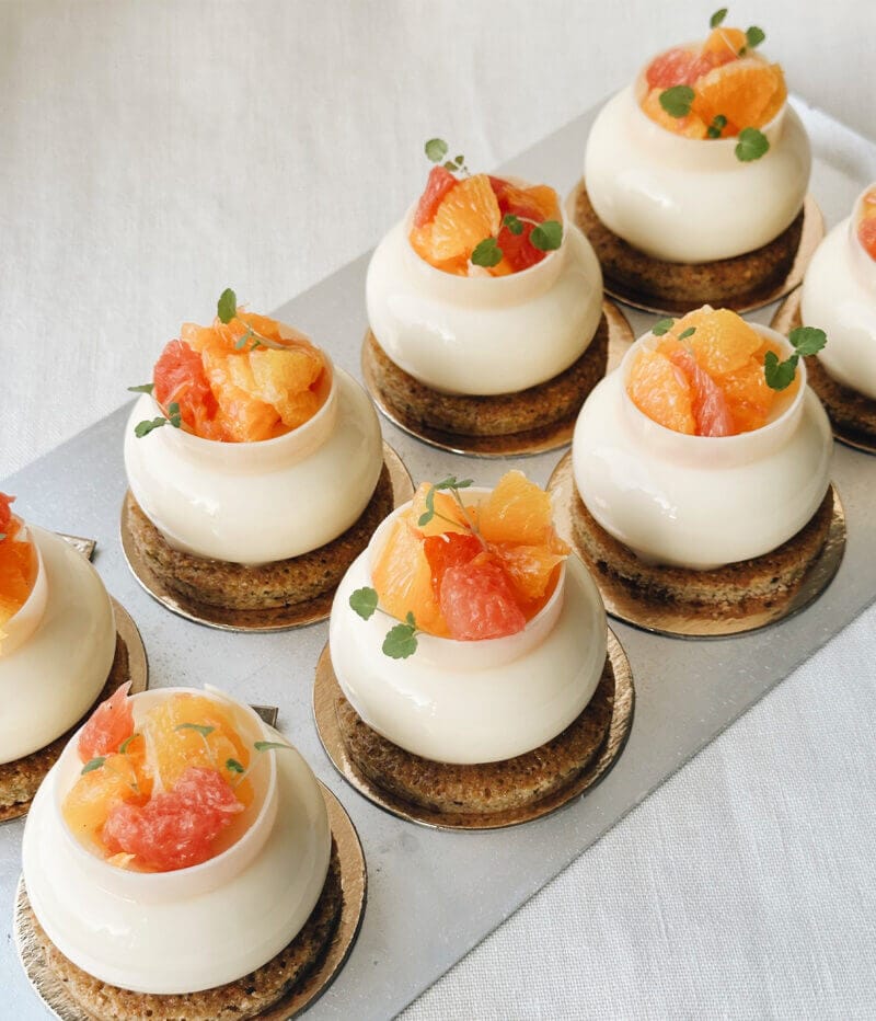 Ett elegant arrangemang av åtta runda bakverk, toppade med livfulla citrusfrukter och färska gröna blad, sitter lockande på en metallbricka - en härlig blandning av smak och presentation inom mat och dryck.