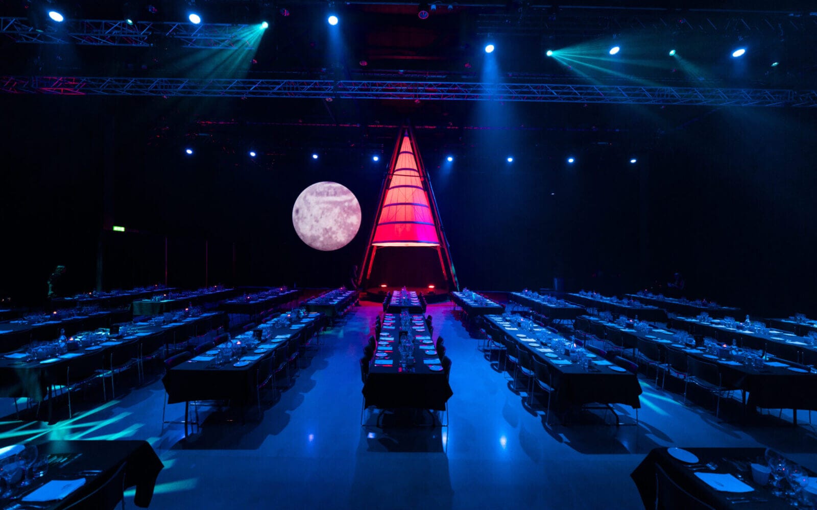 Atmosfären under Arrow Innovation Awards 2024 utspelar sig i ett svagt upplyst evenemangsutrymme med rader av bord förberedda för middagar. En stor, upplyst triangulär struktur pryder scenen mot en månbelyst bakgrund och skapar en fängslande miljö.