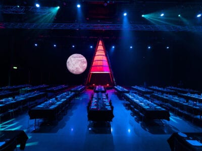 Atmosfären under Arrow Innovation Awards 2024 utspelar sig i ett svagt upplyst evenemangsutrymme med rader av bord förberedda för middagar. En stor, upplyst triangulär struktur pryder scenen mot en månbelyst bakgrund och skapar en fängslande miljö.