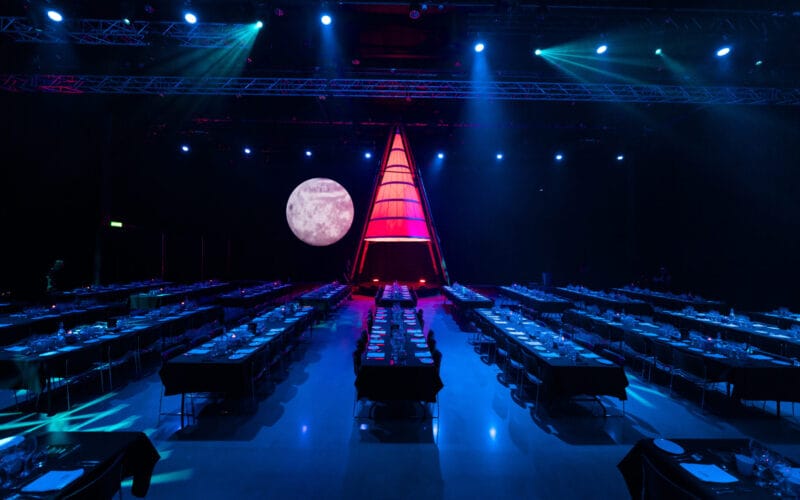 Atmosfären under Arrow Innovation Awards 2024 utspelar sig i ett svagt upplyst evenemangsutrymme med rader av bord förberedda för middagar. En stor, upplyst triangulär struktur pryder scenen mot en månbelyst bakgrund och skapar en fängslande miljö.