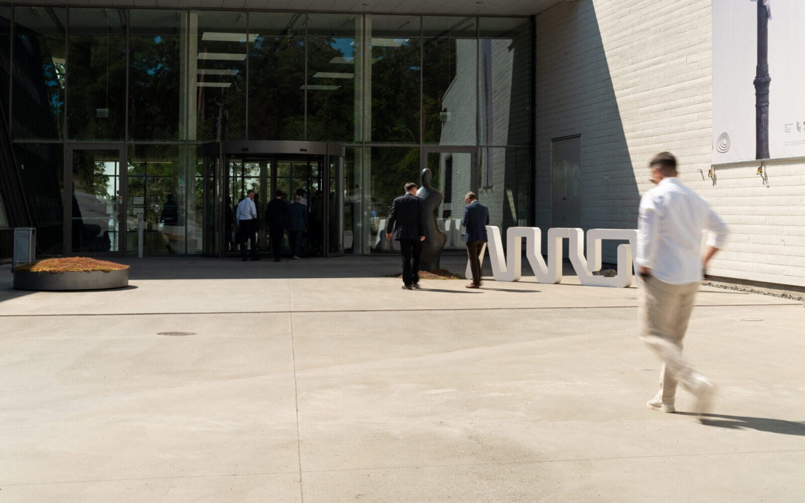 Flera personer, mestadels i affärskläder, går mot en modern byggnadsingång med glasdörrar en solig dag, kanske när de deltar i det prestigefyllda Arrow Innovation Awards 2024. I förgrunden promenerar någon i vardagskläder bort från entrén.