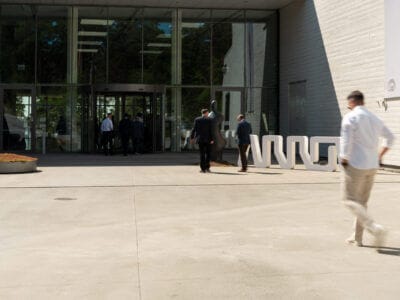 Flera personer, mestadels i affärskläder, går mot en modern byggnadsingång med glasdörrar en solig dag, kanske när de deltar i det prestigefyllda Arrow Innovation Awards 2024. I förgrunden promenerar någon i vardagskläder bort från entrén.