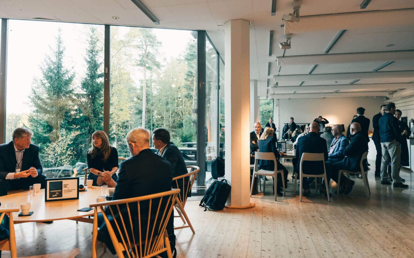 Människor som sitter och samtalar i ett modernt, rymligt rum med stora fönster med utsikt över ett skogsområde.
