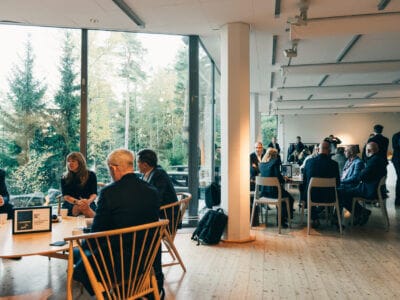 Människor som sitter och samtalar i ett modernt, rymligt rum med stora fönster med utsikt över ett skogsområde.