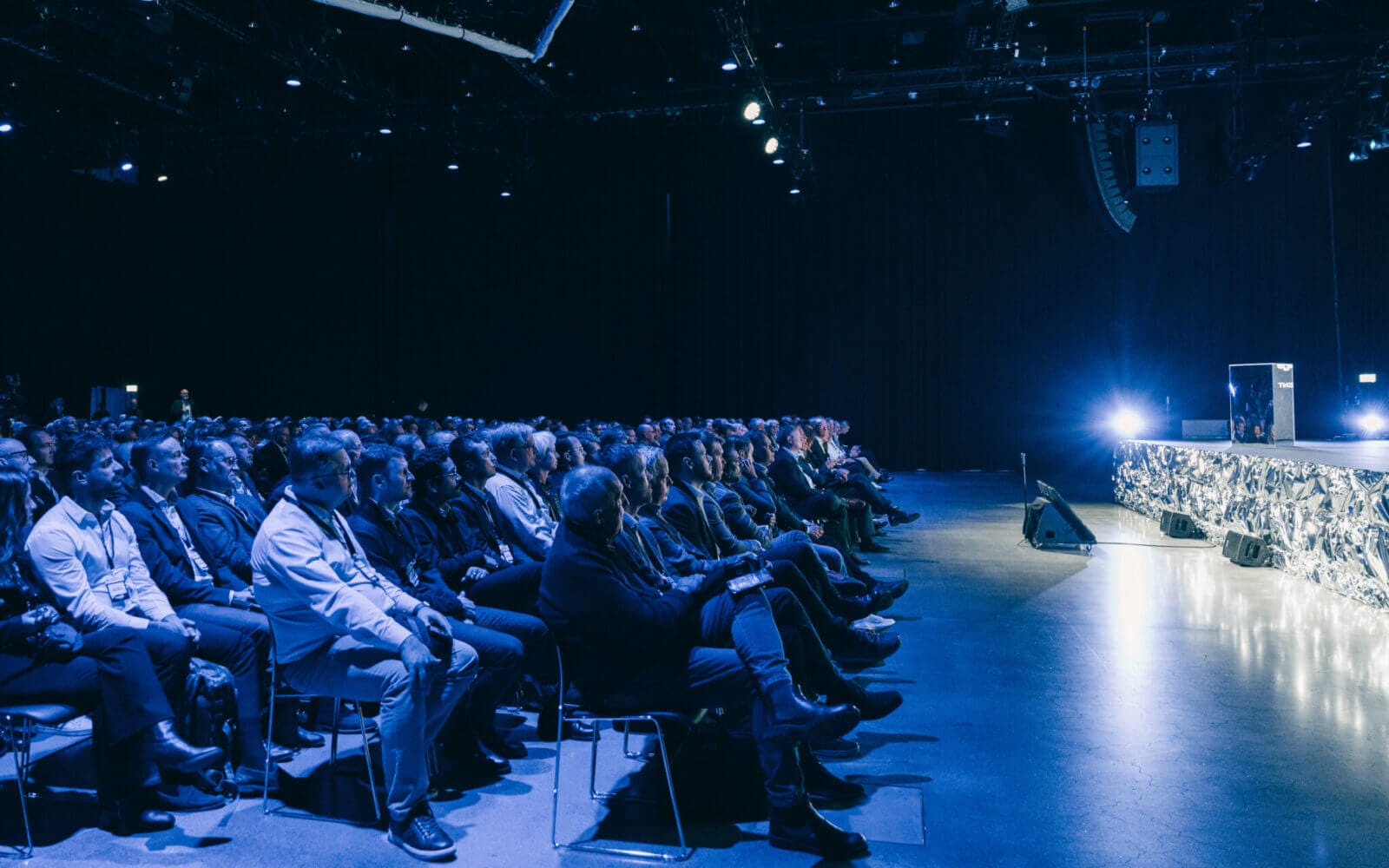 Publiken sitter i en svagt upplyst auditorium, vänd mot en scen med starkt ljus.