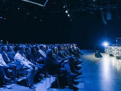 Publiken sitter i en svagt upplyst auditorium, vänd mot en scen med starkt ljus.