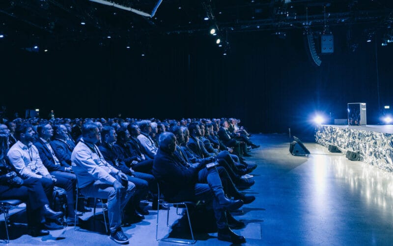 Publiken sitter i en svagt upplyst auditorium, vänd mot en scen med starkt ljus.