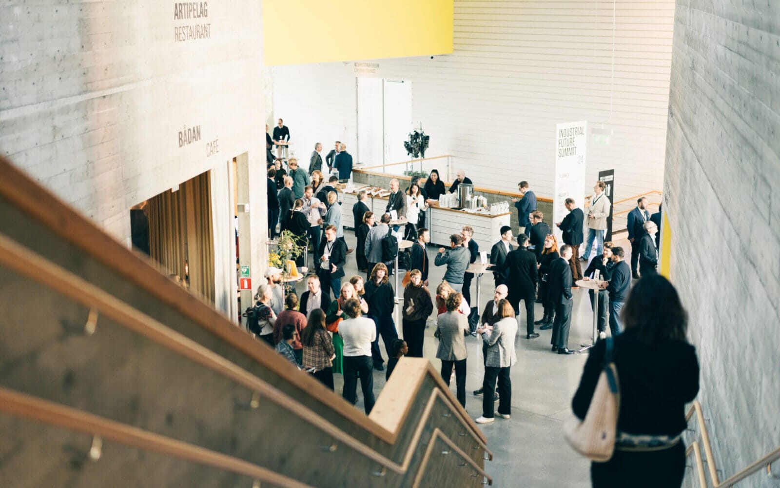 En grupp människor umgås och samlas på en modern inomhuslokal med stora betongväggar och en trappa.
