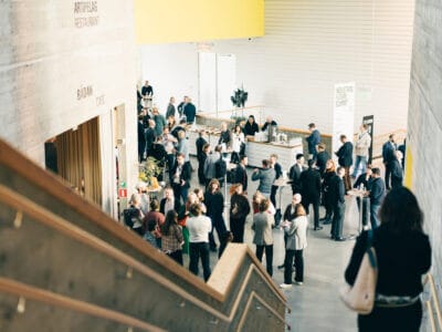 En grupp människor umgås och samlas på en modern inomhuslokal med stora betongväggar och en trappa.