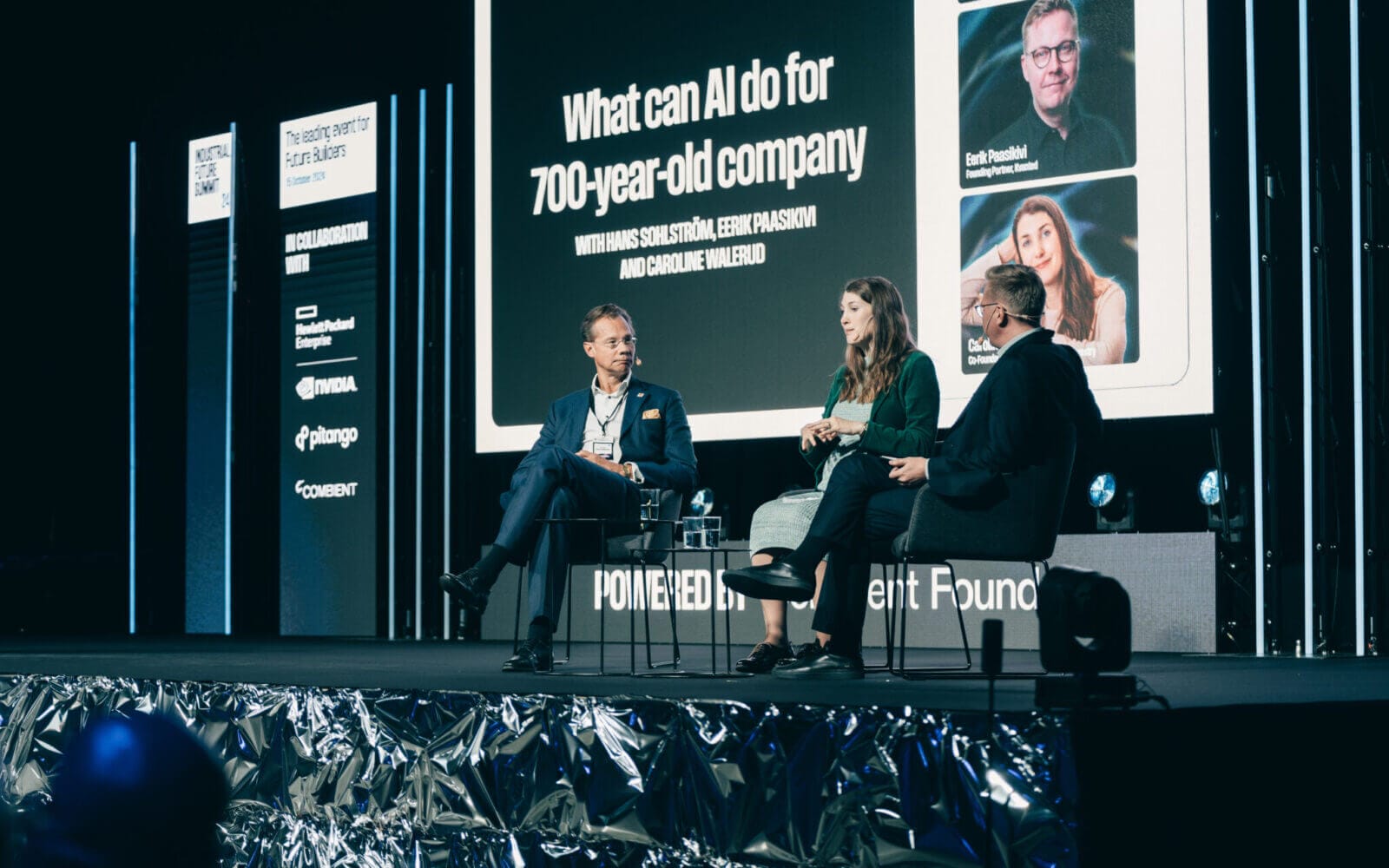 Tre personer satt på scenen och diskuterade "Vad kan AI göra för ett 700-årigt företag" med en bakgrund som visar deltagarnas foton och namn.