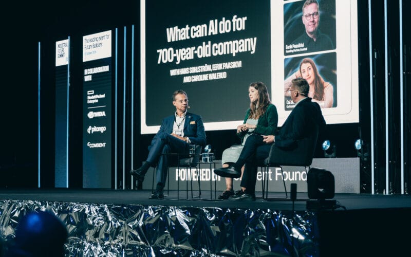 Tre personer satt på scenen och diskuterade "Vad kan AI göra för ett 700-årigt företag" med en bakgrund som visar deltagarnas foton och namn.