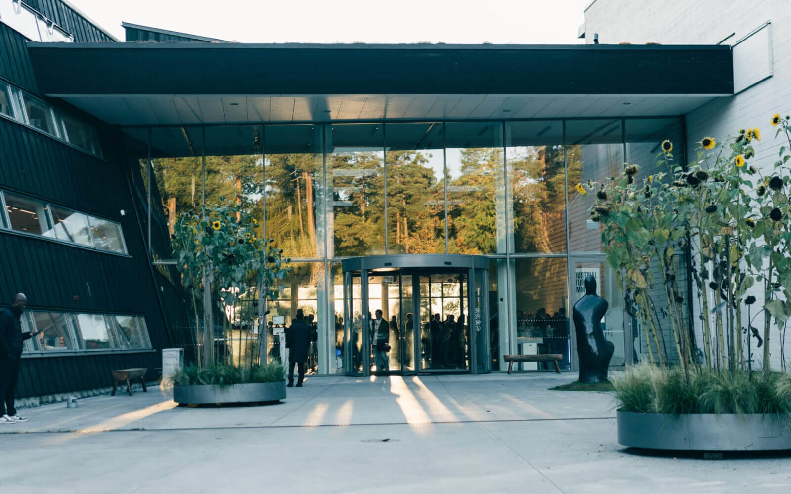 Modern glasbyggnadsingång med svängdörr, flankerad av geometriska väggar och höga krukväxter. Människor är synliga genom glaset och solljus kastar långa skuggor på marken.