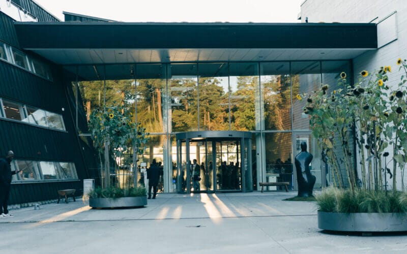 Modern glasbyggnadsingång med svängdörr, flankerad av geometriska väggar och höga krukväxter. Människor är synliga genom glaset och solljus kastar långa skuggor på marken.