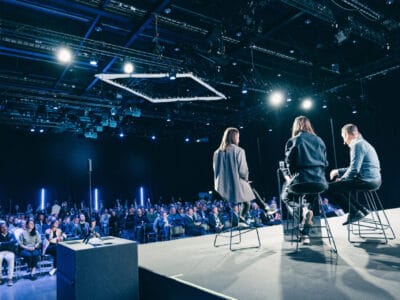 Tre personer på scenen talar till en sittande publik i en konferenssal med blå belysning.