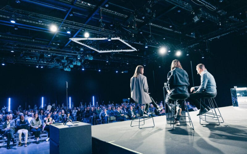 Tre personer på scenen talar till en sittande publik i en konferenssal med blå belysning.