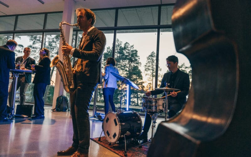 En saxofonist och trummis uppträder inomhus framför ett stort fönster med träd och solnedgång i bakgrunden. Silhouetted människor samtalar vid ett bord till vänster.