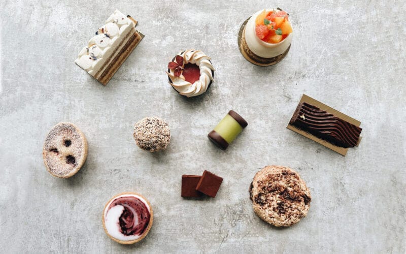 En rad olika bakverk och choklad, konstfullt arrangerade på en strukturerad yta, framkallar den charmiga atmosfären i Bådan Café & Konditori på Värmdö, inbäddat i Stockholms pittoreska skärgård.