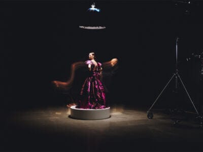 En kvinna i lila klänning står på en rund plattform i en mörk studio och fångar på ett elegant sätt essensen av Lars Wallin Atelier – 35 Years of Fashion Stories. Rörelseoskärpa effekter förbättrar scenen, med studioljus och utrustning delvis synliga.