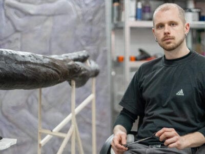 En man i svart t-shirt sitter bredvid ett stort, mörkt skulpturalt konstverk i en ateljé med konstnärsmaterial och en målning i bakgrunden, möjligen under förberedelse inför Fredrik Roos stipendium 2025.