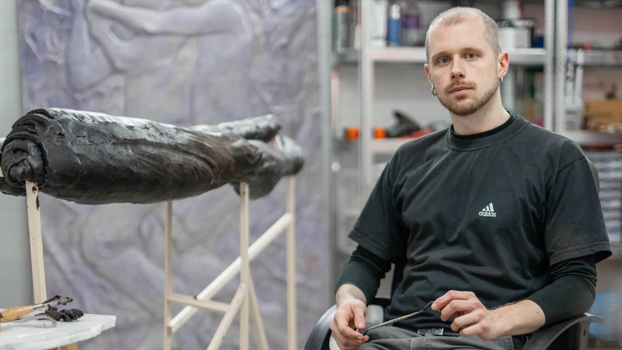 En man i svart t-shirt sitter bredvid ett stort, mörkt skulpturalt konstverk i en ateljé med konstnärsmaterial och en målning i bakgrunden, möjligen under förberedelse inför Fredrik Roos stipendium 2025.